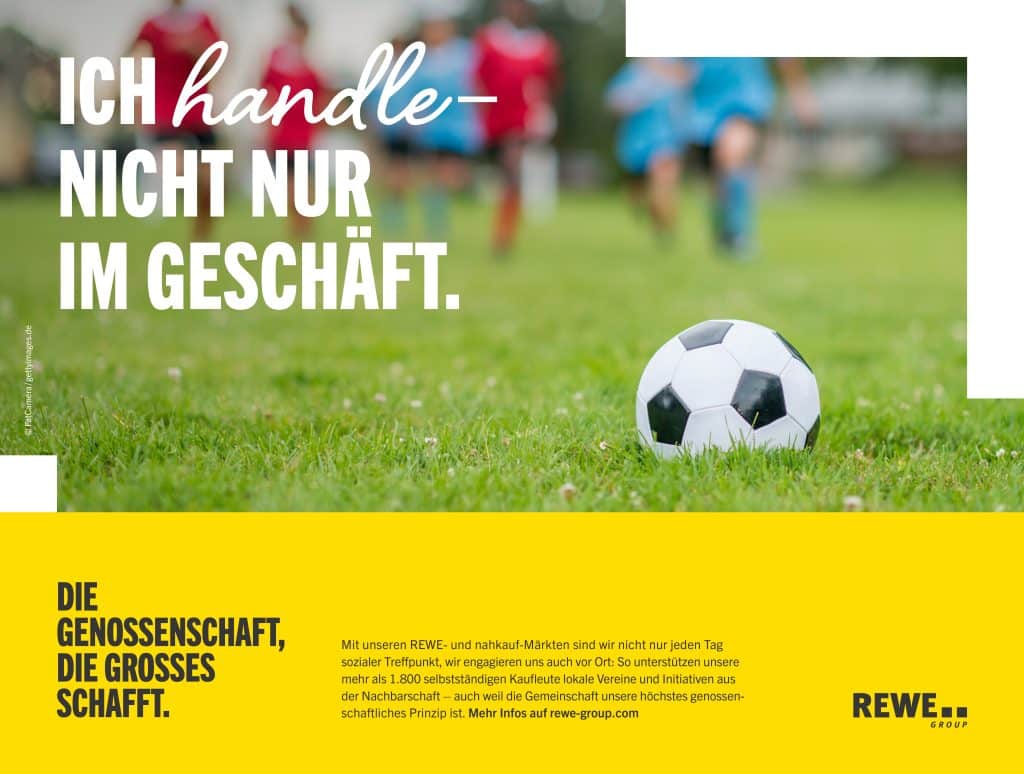 Auf einer Rasenfläche liegt ein Fußball im Vordergrund. Im Hintergrund sind mehrere spielende Jugendliche.
