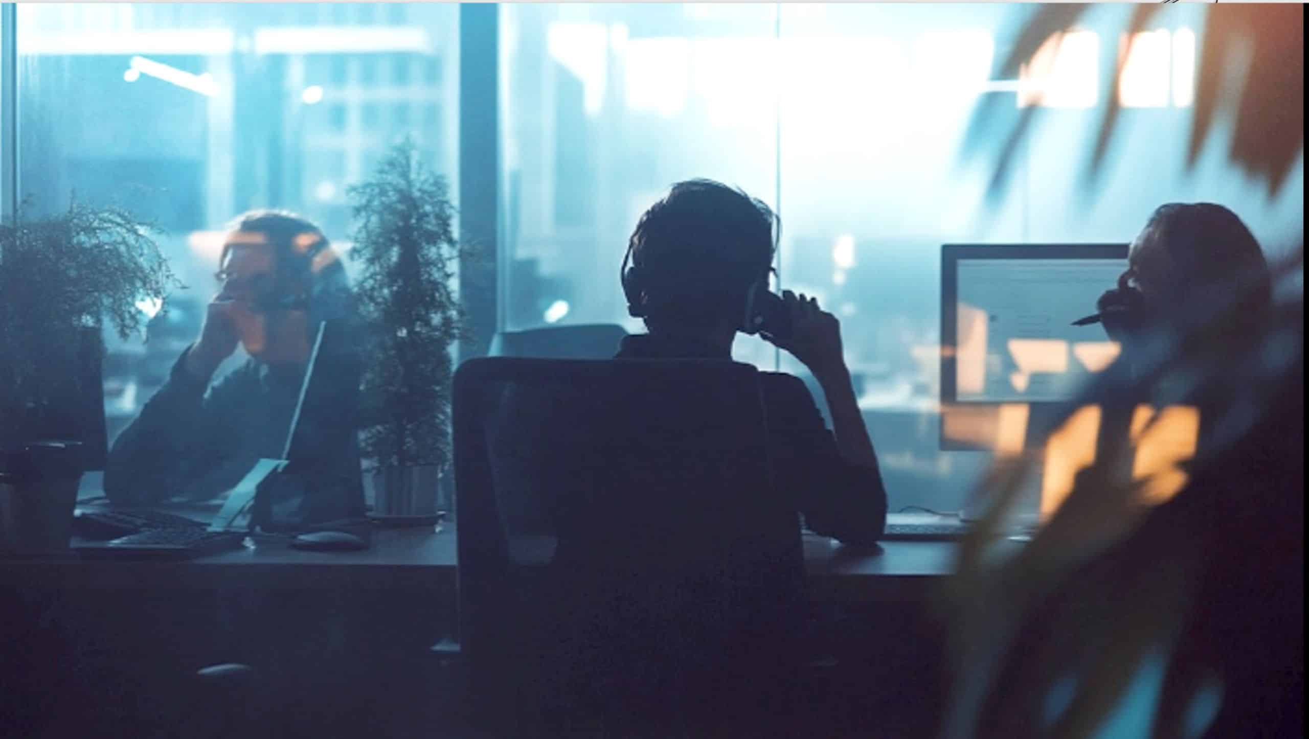 In einem Büro sitzen mehrere Personen mit Headsets an Schreibtischen und arbeiten an Computern. Die Szene ist in gedämpftem Licht gehalten, während Pflanzen und Glaswände den Raum strukturieren.
