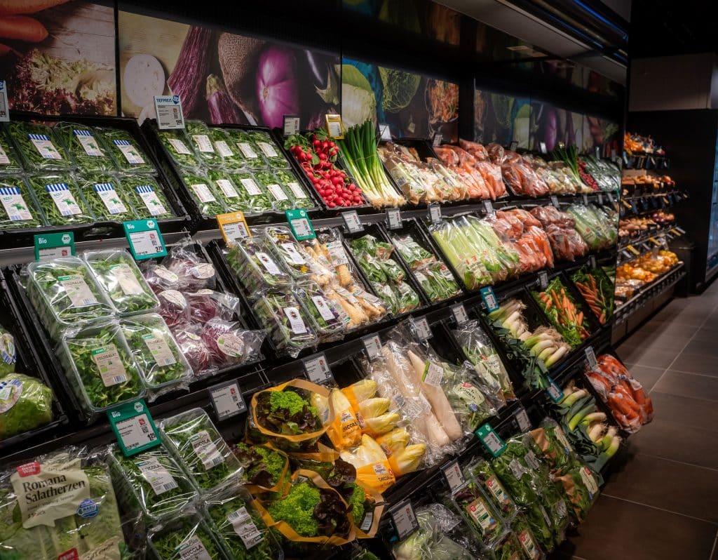The picture shows a supermarket shelf with a large selection of fresh vegetables, including lettuce, herbs, leeks, carrots and packaged leafy vegetables.