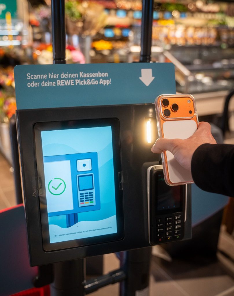 The picture shows a station in the supermarket where a smartphone is held in front of a scanner to scan a receipt or the REWE Pick&Go app.