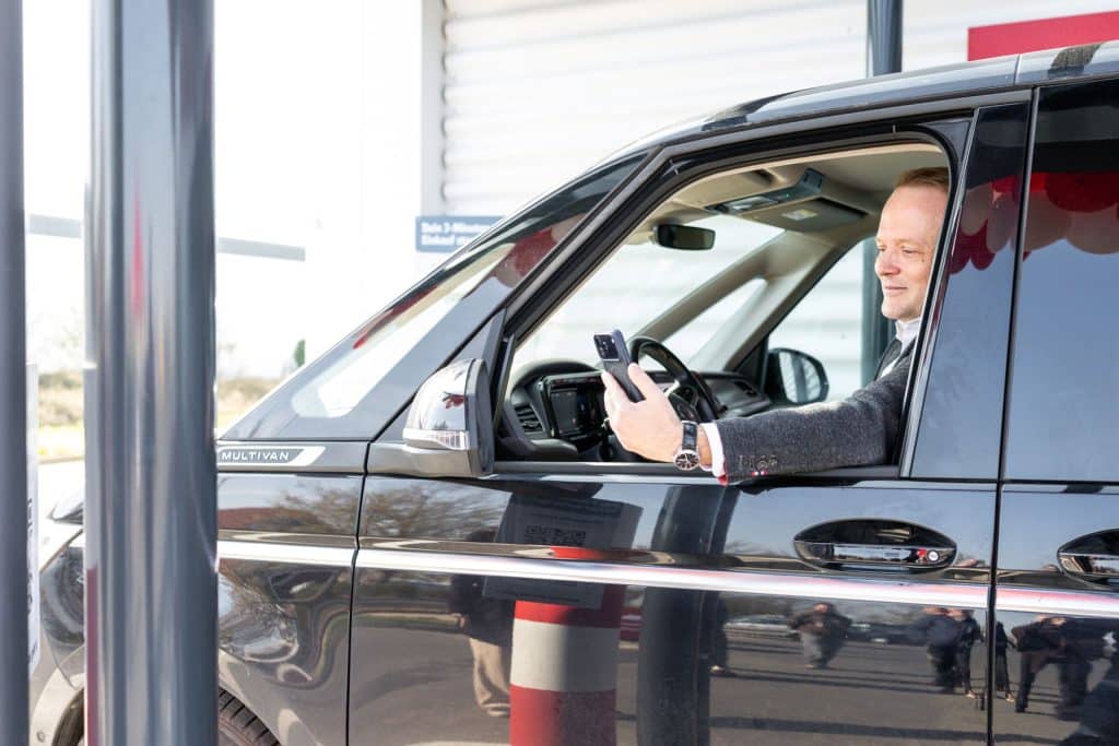 Eine Person sitzt in einem Fahrzeug auf dem Fahrersitz und hält ein Smartphone aus dem geöffneten Seitenfenster.