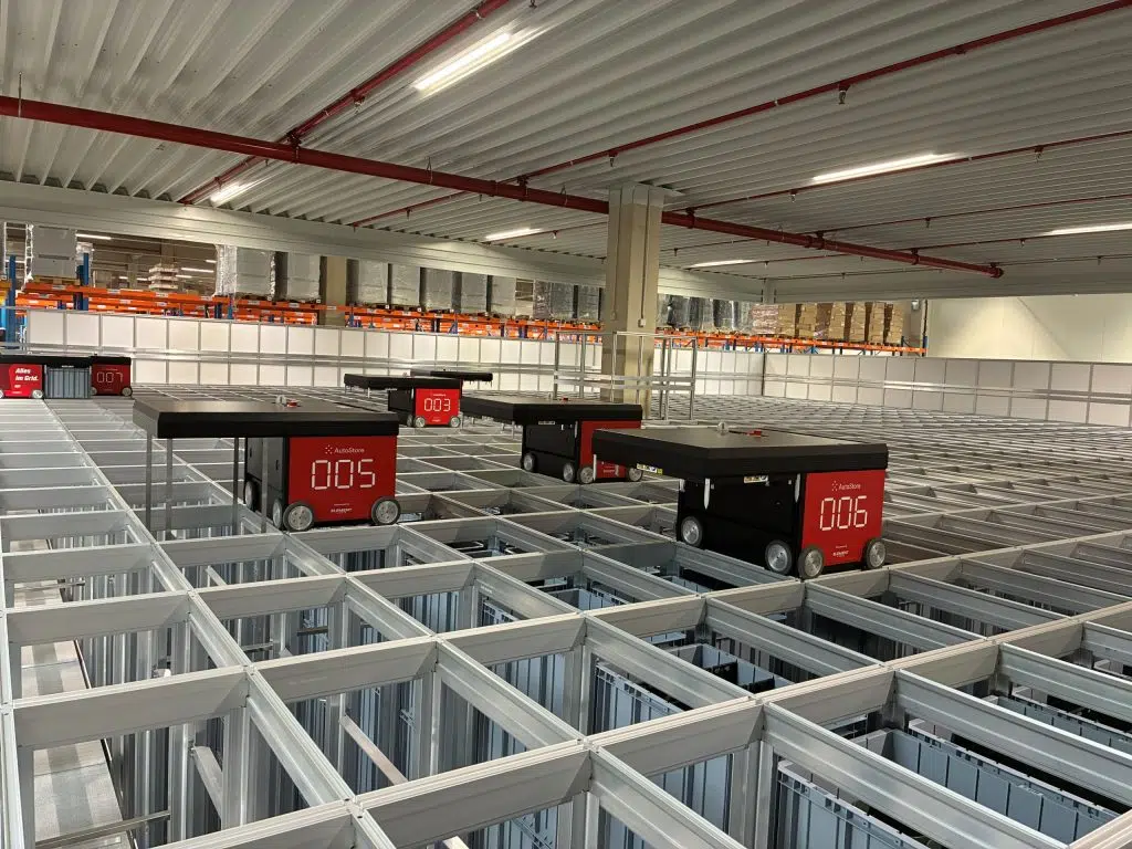 Automatisiertes Lagersystem mit mehreren kleinen robotischen Transportfahrzeugen, die auf einem rasterförmigen Metallgestell in einer Lagerhalle Waren bewegen.