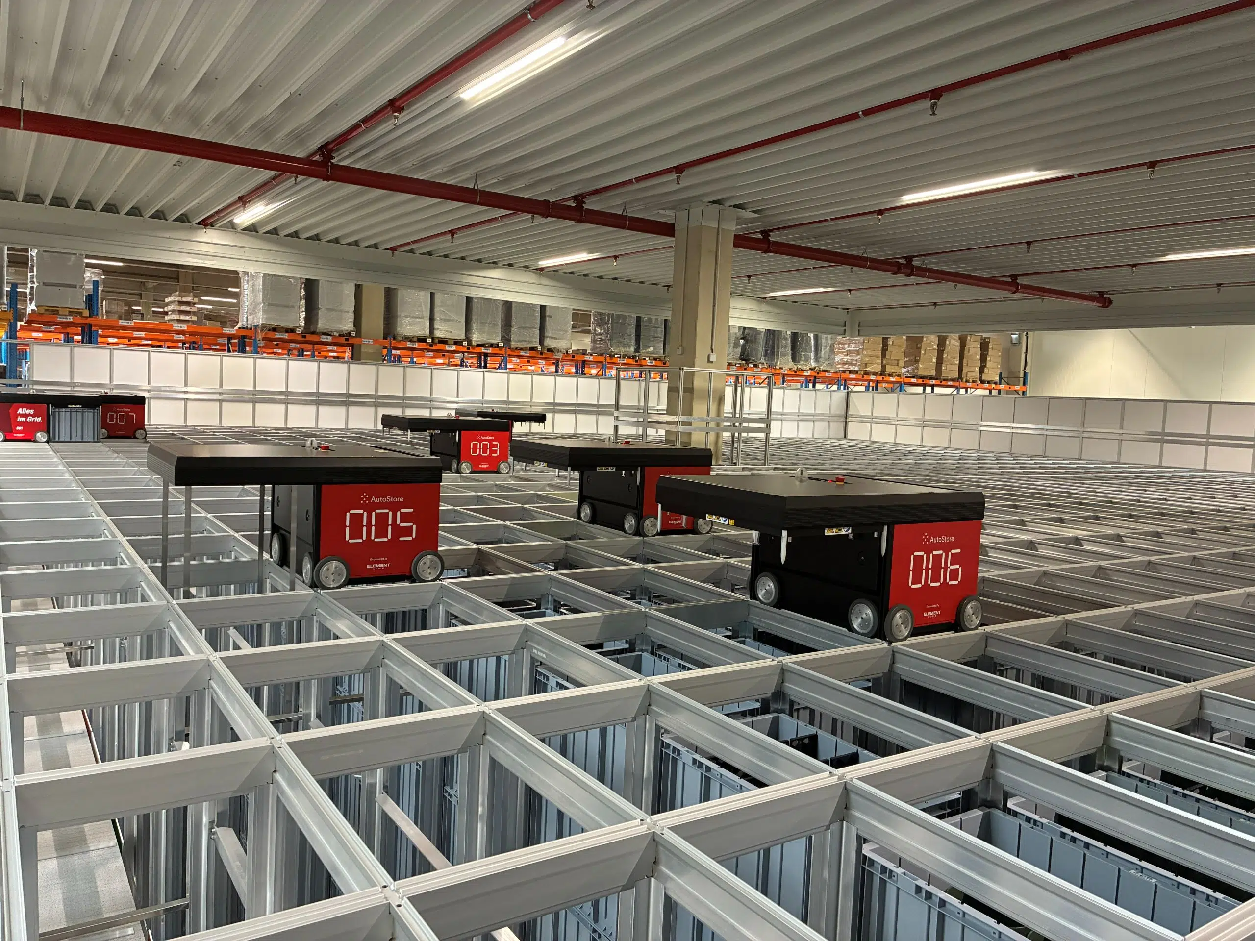 Automatisiertes Lagersystem mit mehreren kleinen robotischen Transportfahrzeugen, die auf einem rasterförmigen Metallgestell in einer Lagerhalle Waren bewegen.