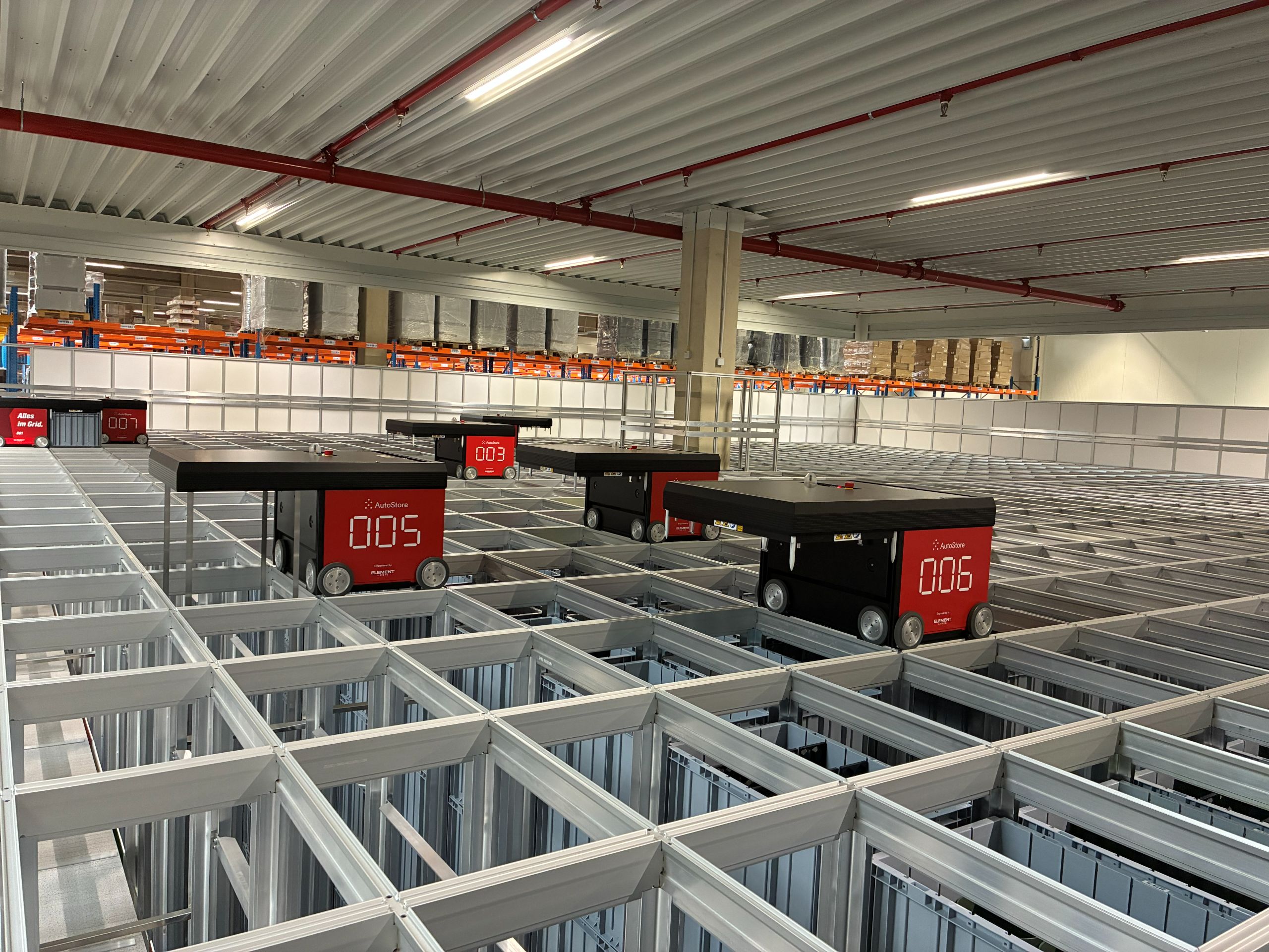 Automated storage system with several small robotic transport vehicles that move goods on a grid-shaped metal frame in a warehouse.