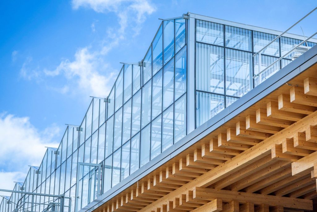 Eine moderne, gläserne Gewächshauskonstruktion steht auf dem Dach eines Gebäudes mit sichtbarer Holzkonstruktion vor blauem Himmel mit Wolken.