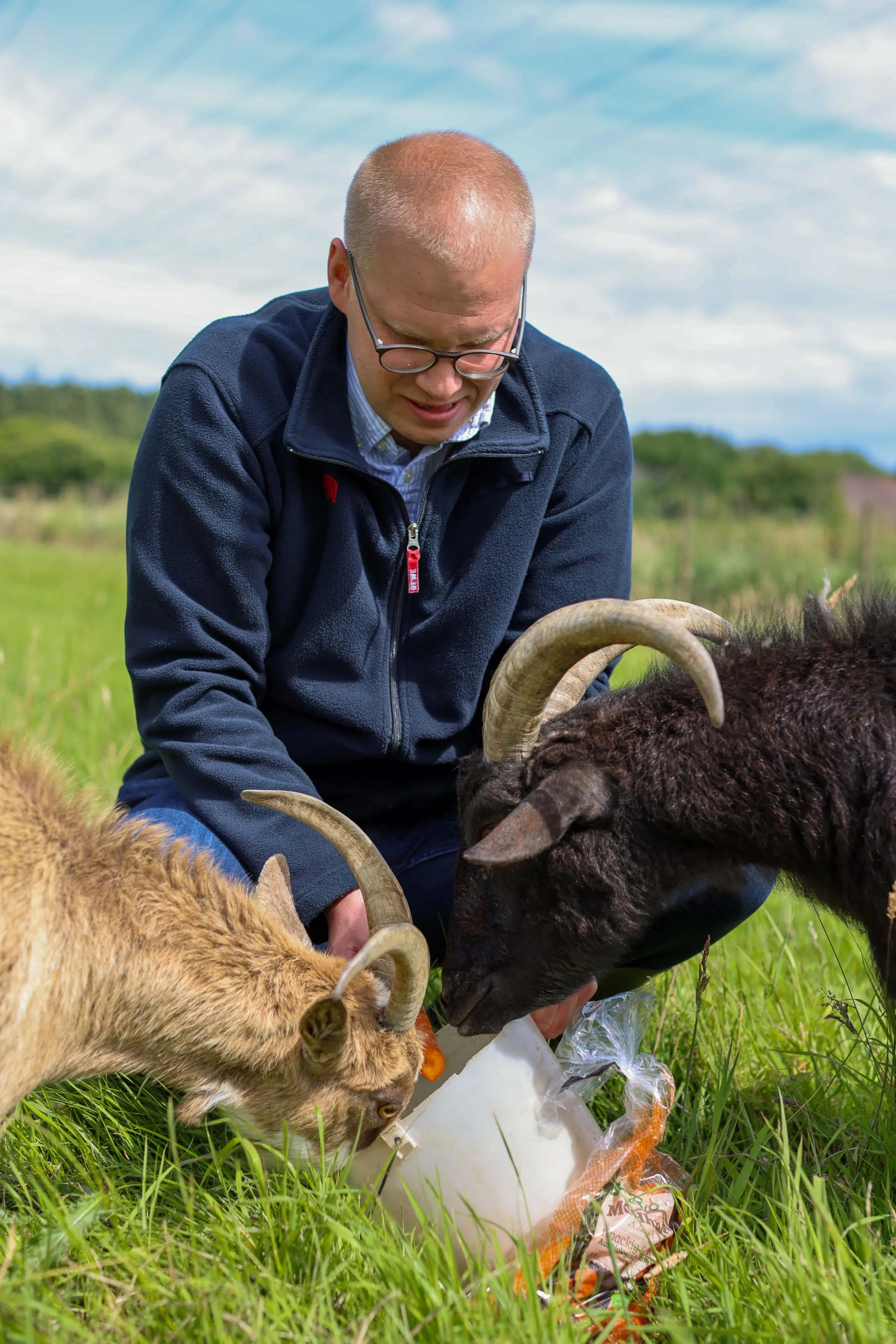 REWE-Kaufmann, Simon Bacher füttert zwei gehörnte Ziegen auf einer grünen Wiese mit Karotten aus einer Futterschale.