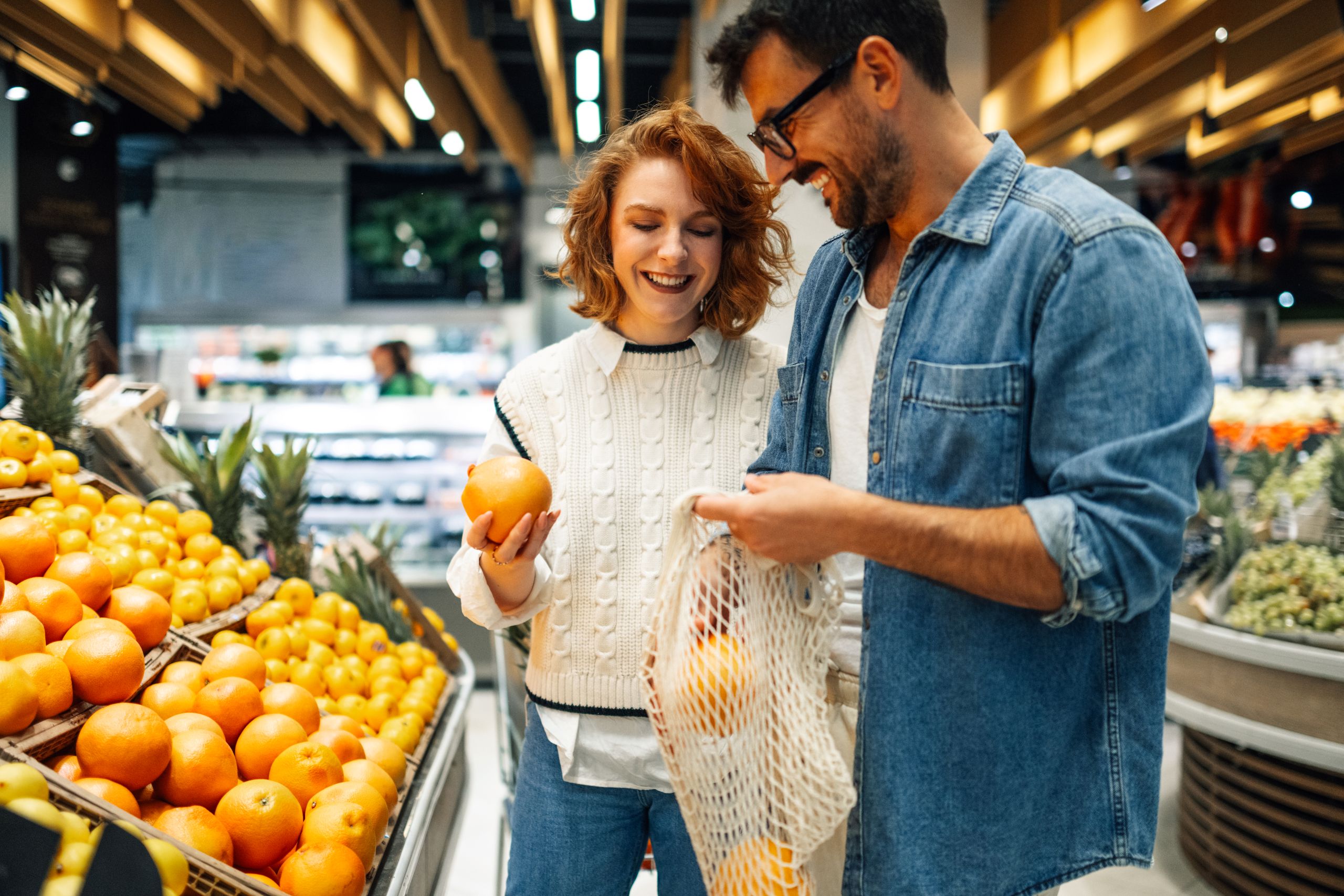 Ein Paar steht in einem Supermarkt vor einer Auslage mit Orangen und legen Obst in ein Netz.