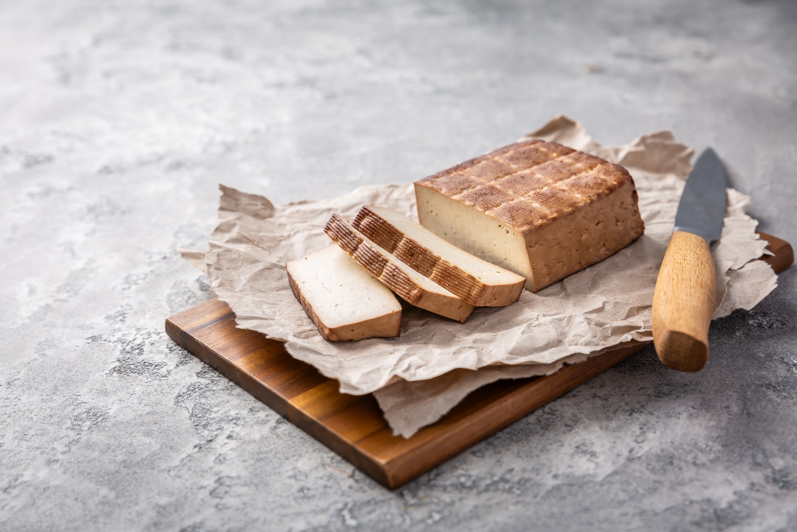 Ein Block geräucherter Tofu mit mehreren abgeschnittenen Scheiben liegt auf Backpapier auf einem Holzbrett, daneben befindet sich ein Messer mit Holzgriff.