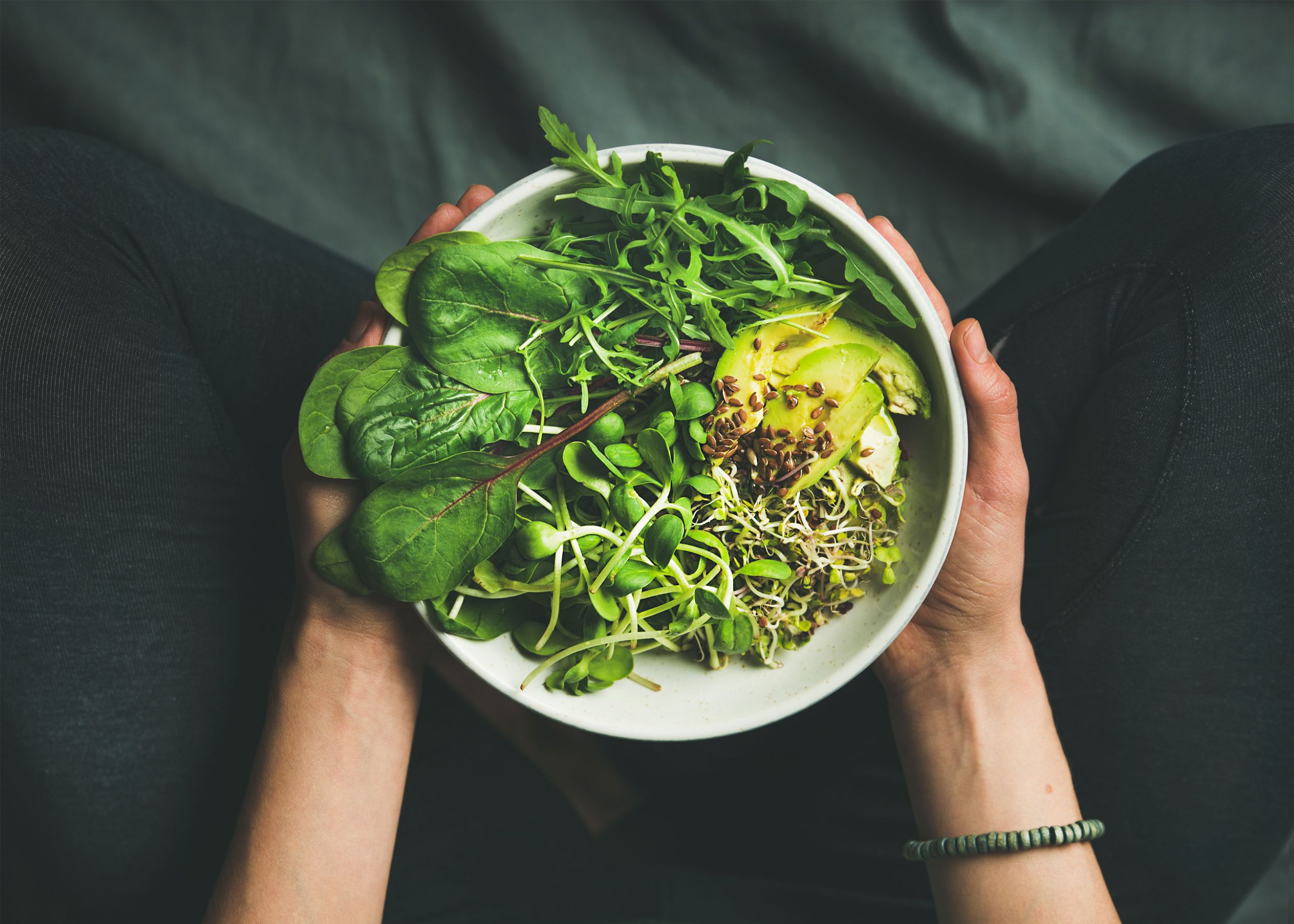 Schale mit frischem grünen Salat aus Spinat, Rucola, Sprossen und Avocado, gehalten in zwei Händen.