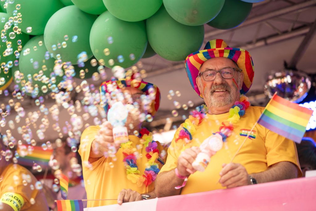 Teilnehmende auf einem REWE-Pride-Truck mit Regenbogenflaggen, bunten Accessoires und Seifenblasen bei einer CSD-Demonstration.