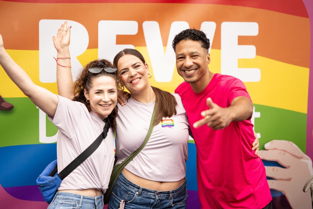 Drei Teilnehmende einer Pride-Veranstaltung posieren vor einer großen Regenbogenwand mit dem Schriftzug „REWE Dein Markt“.