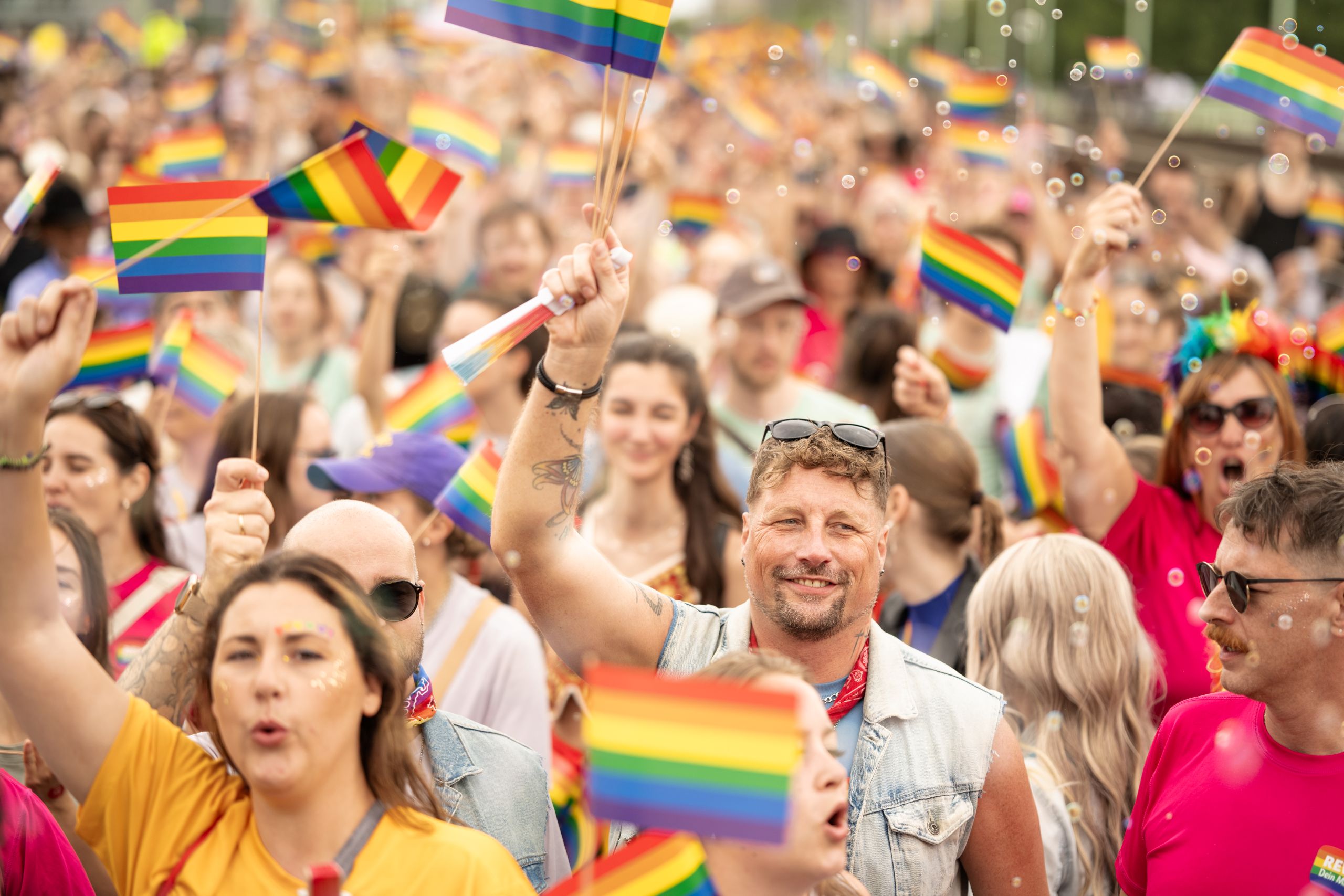 Participants in a Pride march carrying numerous rainbow flags amid a densely packed crowd.