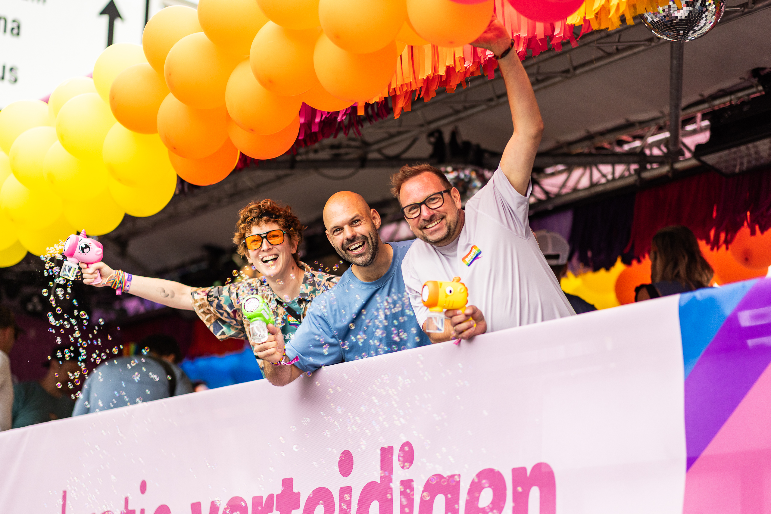 Drei Personen stehen auf einem bunt dekorierten Paradewagen unter gelben Luftballons, halten Seifenblasenpistolen in die Kamera und werden von schwebenden Seifenblasen sowie Regenbogenfarben umgeben.