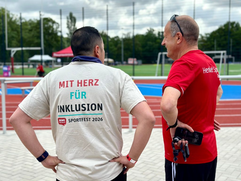 Zwei Personen stehen an einer Leichtathletik- und Sportanlage und blicken auf das Veranstaltungsgelände. Auf dem T-Shirt einer Person steht „Mit Herz für Inklusion – Sportfest 2026“.