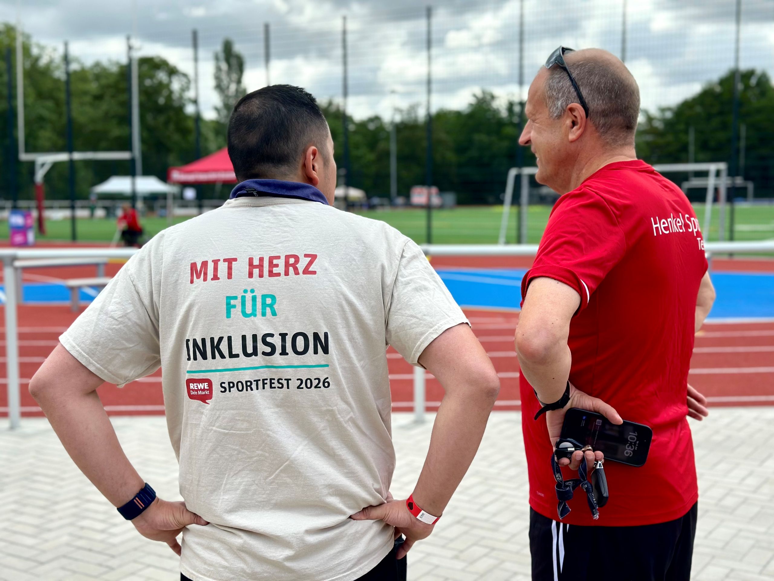 Two people are standing at an athletics and sports complex, looking out at the event grounds. One person’s T-shirt reads “With Heart for Inclusion – Sports Festival 2026.”