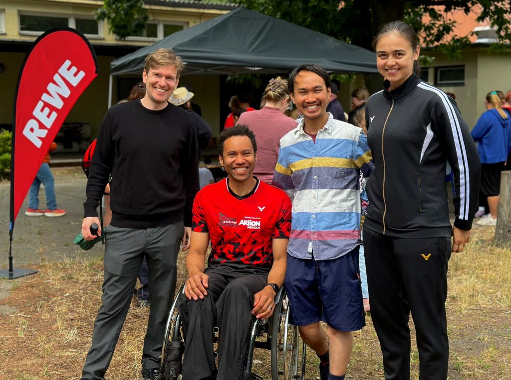 Vier Vertreterinnen und Vertreter des Badmintonsports stehen und sitzen gemeinsam vor einem REWE-Stand auf dem inklusiven Familiensportfest in Berlin-Reinickendorf.