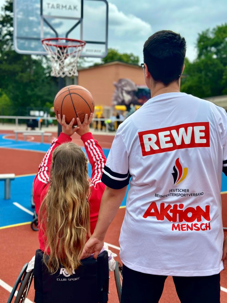 A child is shooting a basketball on a sports field, while a person wearing a T-shirt with the REWE, German Disabled Sports Association, and Aktion Mensch logos stands nearby.