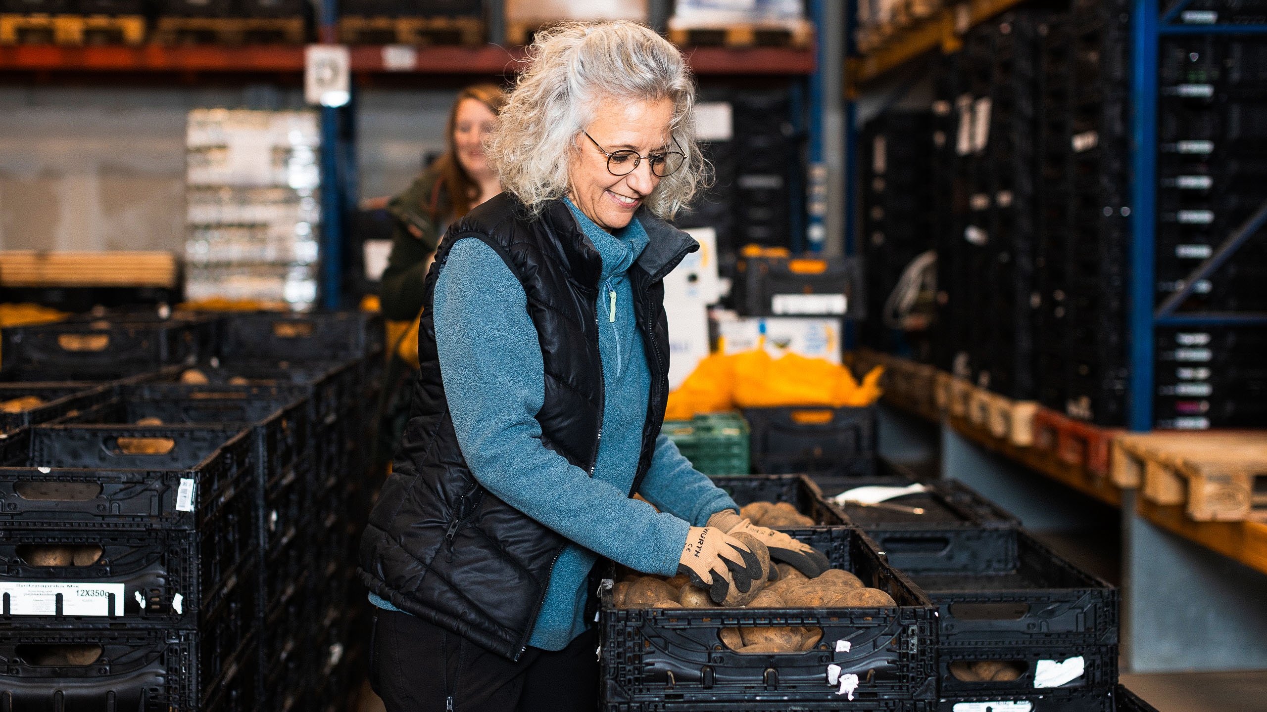 Person mit Arbeitshandschuhen sortiert Kartoffeln in schwarzen Kisten in einer Lagerhalle.