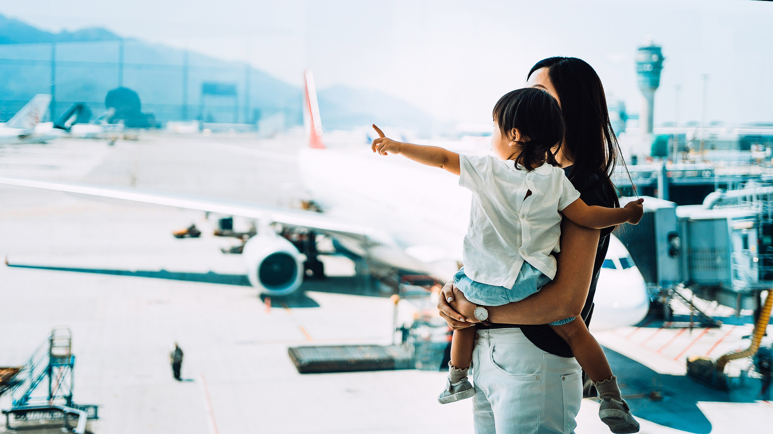 A person is holding a child in their arms in front of a large window at the airport, while the child points to an airplane on the tarmac.
