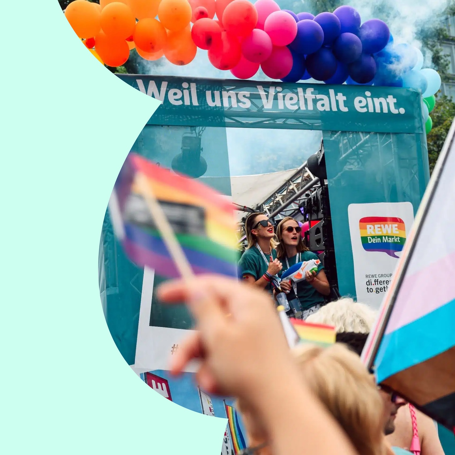 Pride-Veranstaltung mit Regenbogen- und Trans-Pride-Flaggen vor einer Bühne der REWE Group. Über der Bühne steht „Weil uns Vielfalt eint.“, darüber befinden sich bunte Luftballons.