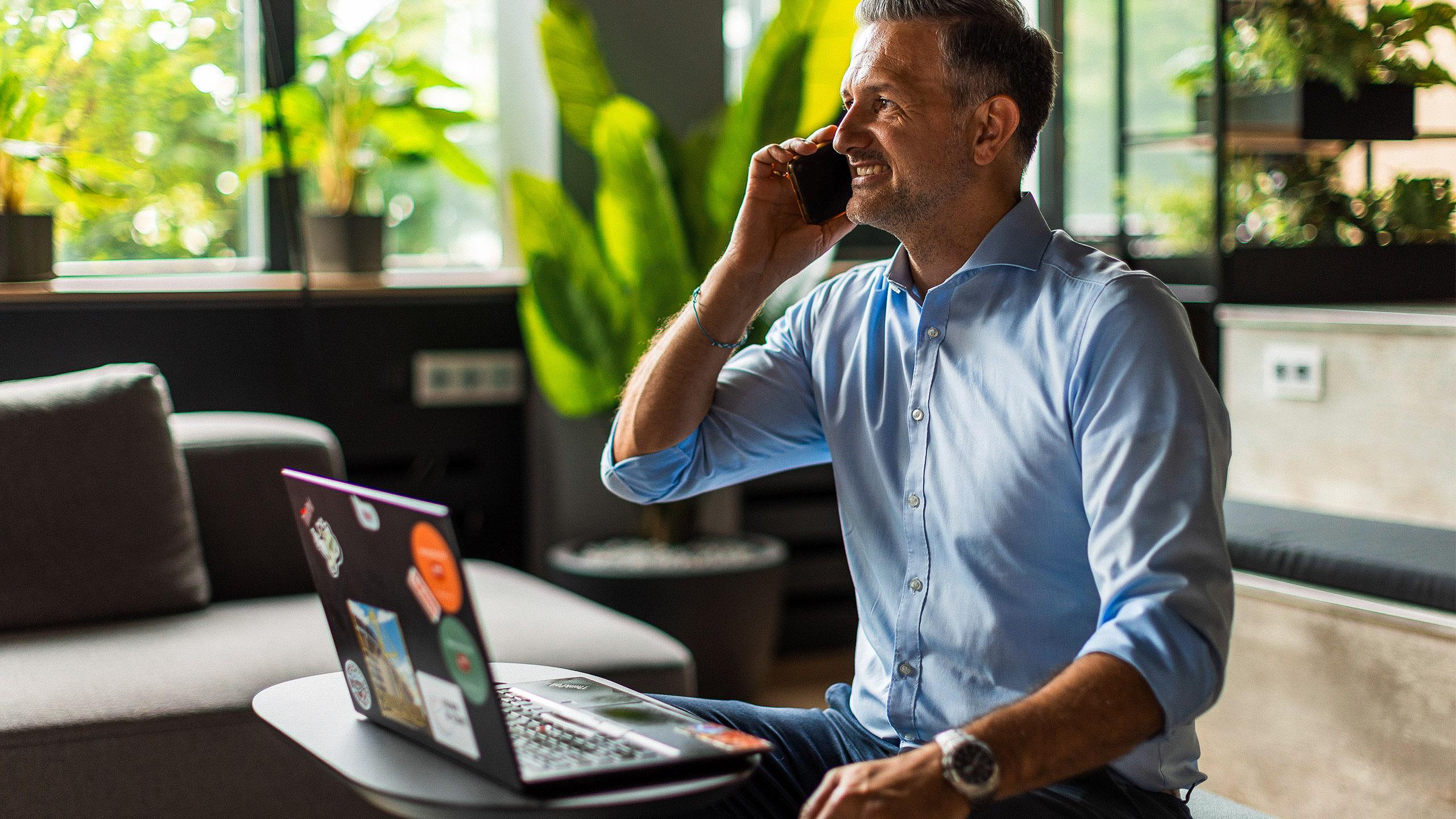 A person is sitting at a table with a laptop and talking on a smartphone in a green office setting.
