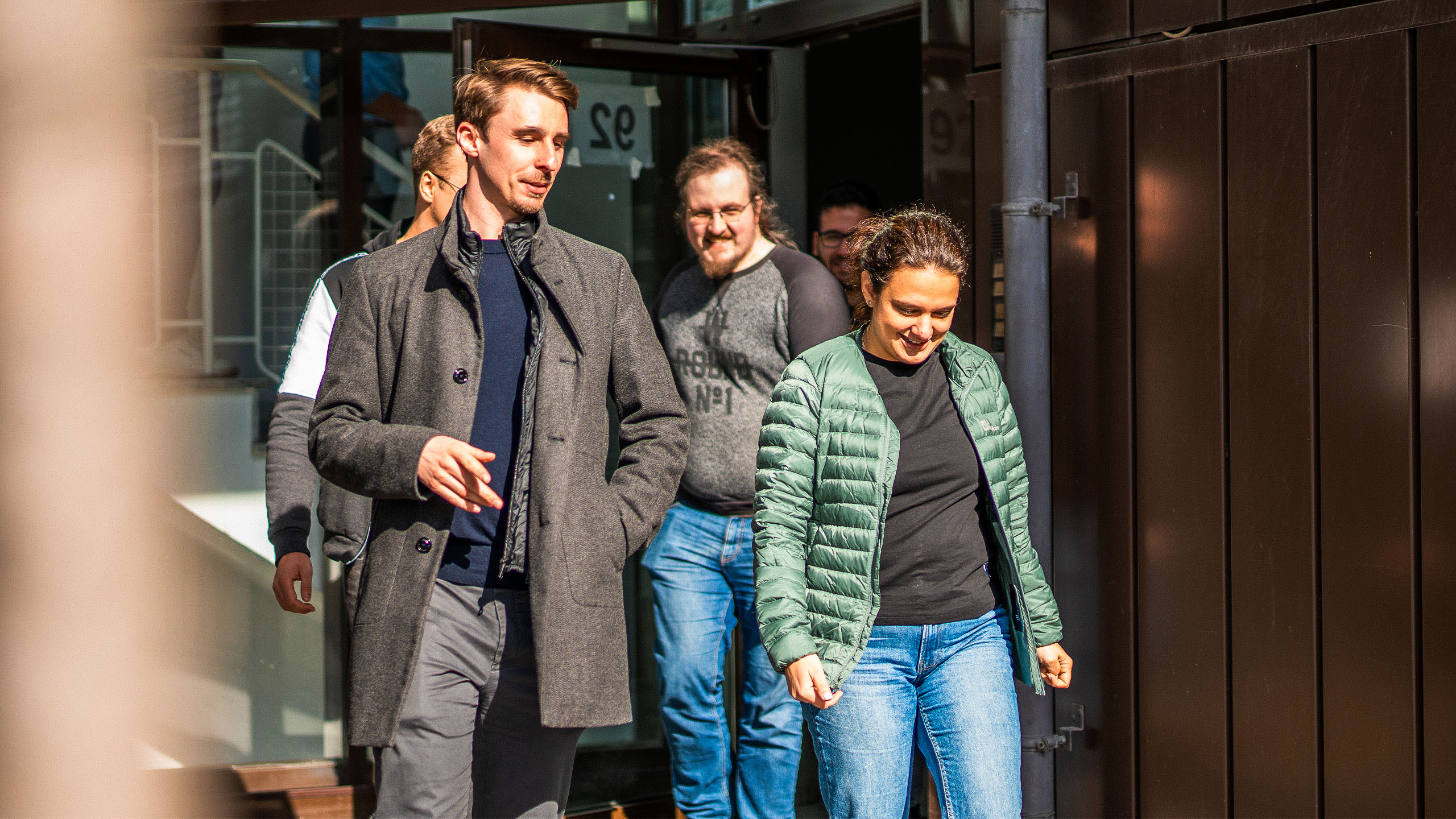 Mehrere Personen verlassen gemeinsam ein Gebäude. Zwei Personen gehen nebeneinander im Vordergrund, weitere folgen dahinter.