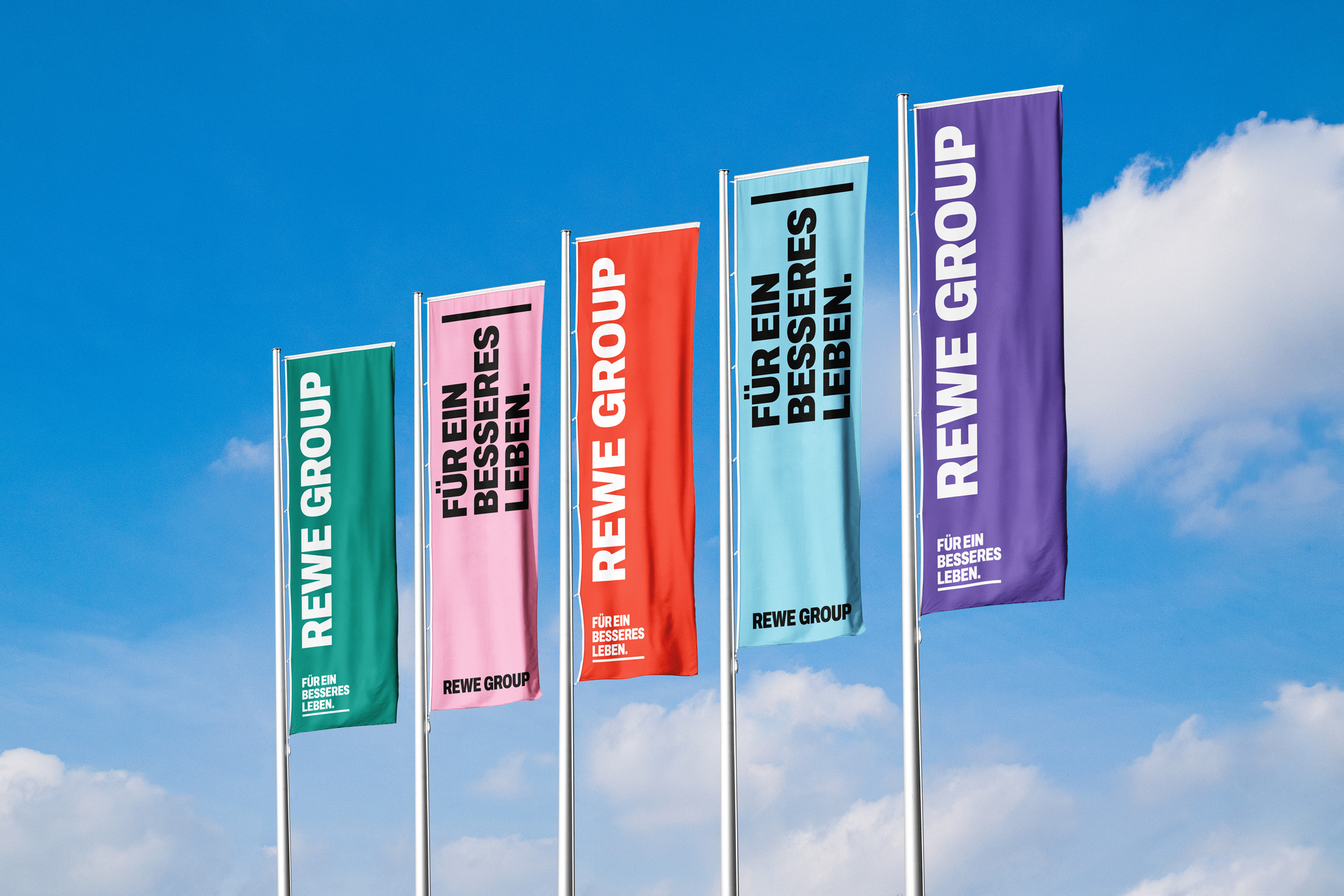 Fünf Flaggen der REWE GROUP in Grün, Rosa, Rot, Hellblau und Violett vor blauem Himmel. Auf den Fahnen stehen „REWE GROUP“ und „Für ein besseres Leben“.