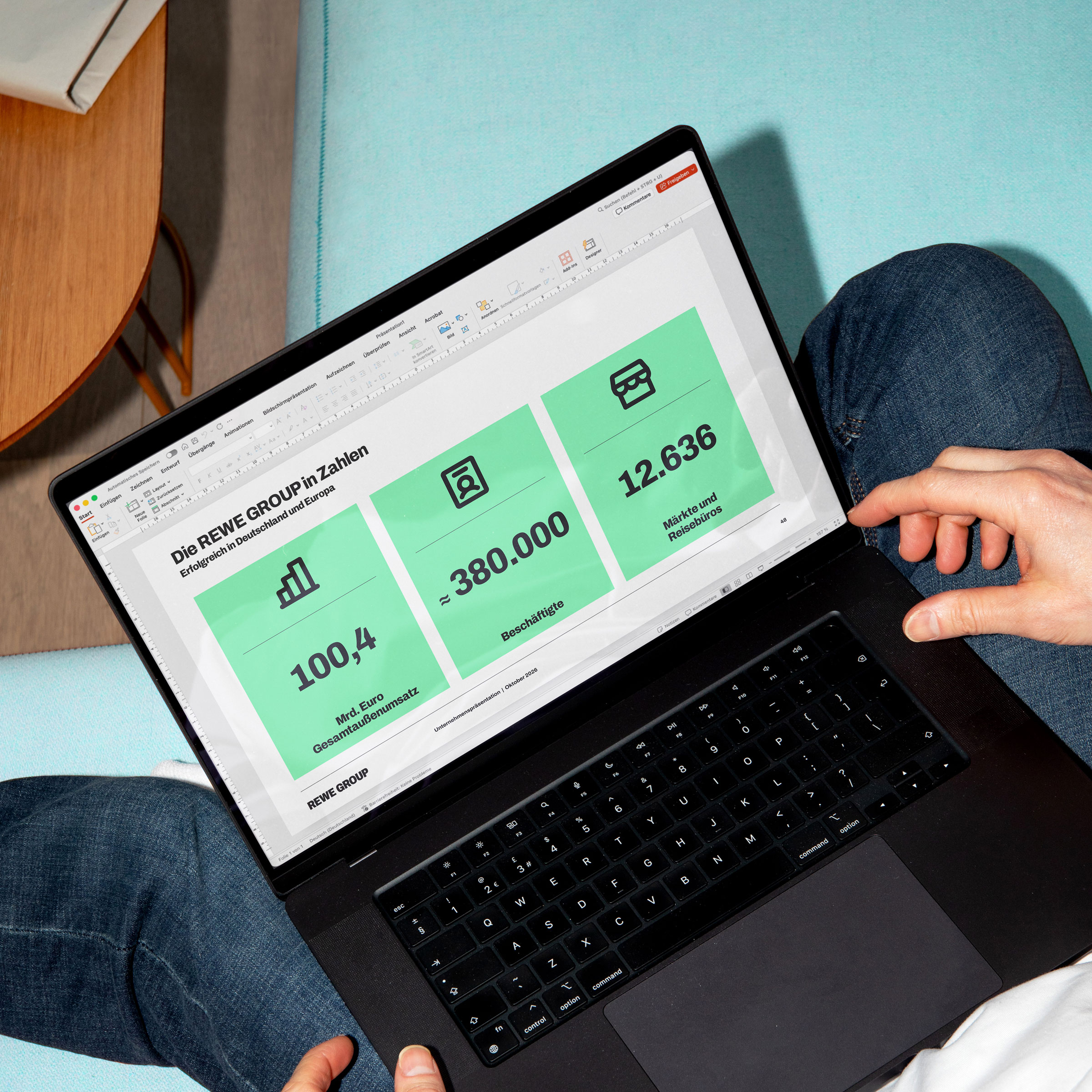 Person mit Laptop auf dem Schoß. Auf dem Bildschirm ist eine Präsentation mit REWE Group Kennzahlen zu sehen: 100,4 Milliarden Euro Gesamtaußenumsatz, rund 380.000 Beschäftigte sowie 12.636 Märkte und Reisebüros.