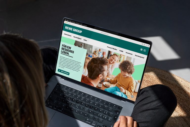 Person mit Laptop auf dem Schoß. Auf dem Bildschirm ist die Website der REWE Group mit dem Schriftzug „Für ein besseres Leben.“ und einem Bild von Personen an einem Tisch geöffnet.