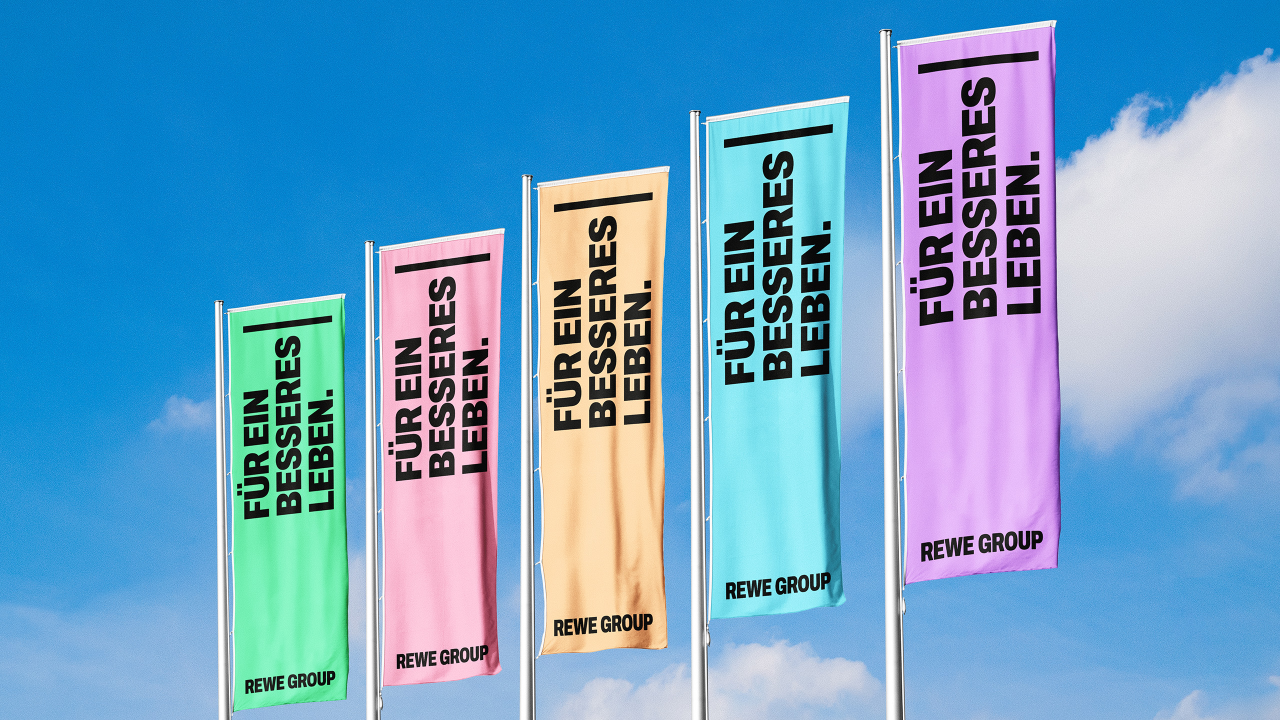 Fünf Fahnen der REWE GROUP in Grün, Rosa, Beige, Hellblau und Violett vor blauem Himmel. Auf den Fahnen steht „Für ein besseres Leben“