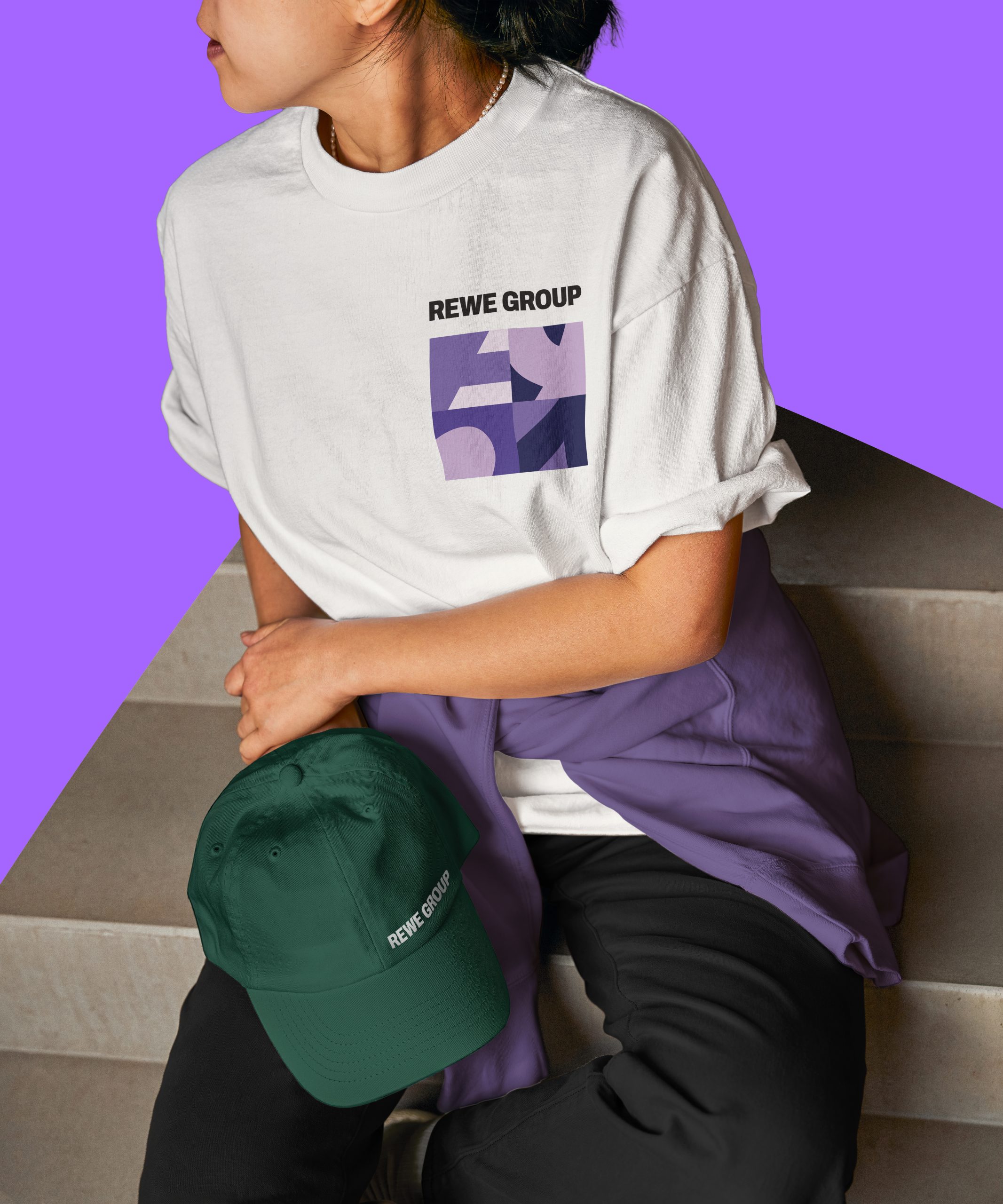 Person sitzt auf einer Treppe und trägt ein weißes T-Shirt mit REWE Group Schriftzug und violettem Grafikmotiv. Auf dem Schoß liegt eine grüne Kappe mit REWE Group Schriftzug.