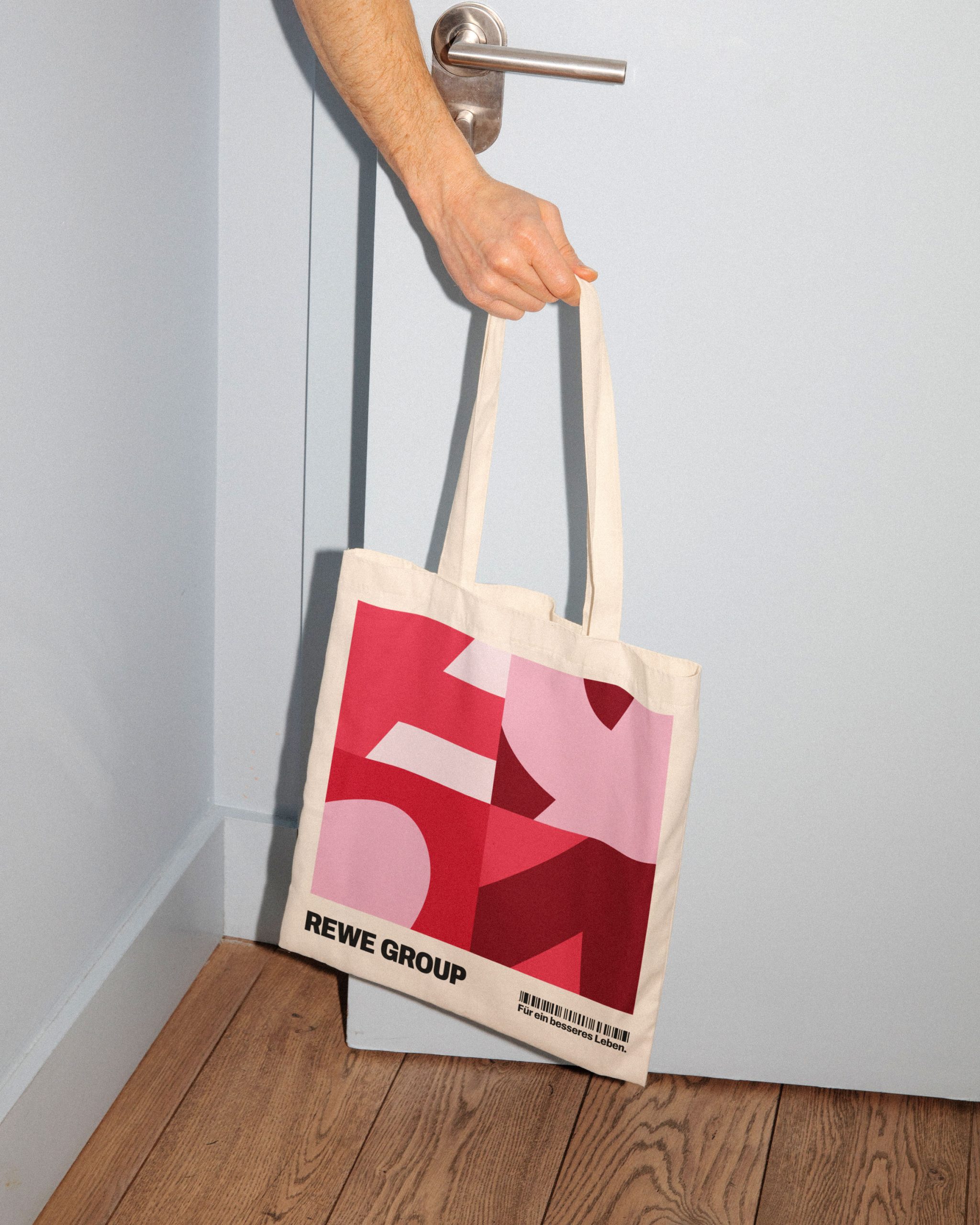 Person hält eine Stofftragetasche mit rot-rosa grafischem Muster und REWE GROUP Schriftzug vor einer weißen Tür.
