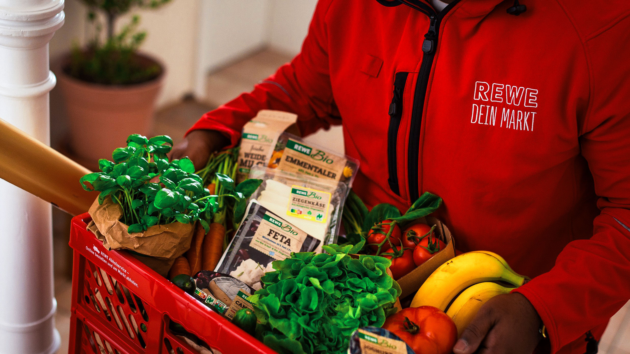 Eine Person in roter Arbeitskleidung trägt eine rote Kiste voller frischer Lebensmittel wie Obst, Gemüse, Salat und Kräuter in einem Treppenhaus.