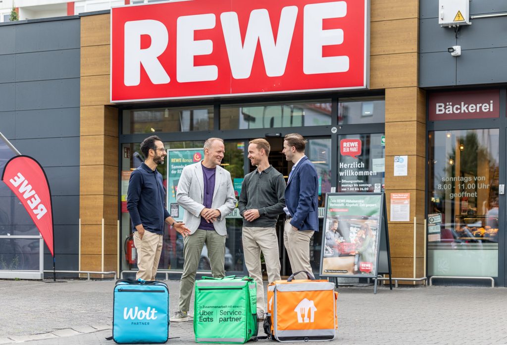 Vier Personen stehen vor dem Eingang eines REWE-Markts. Im Vordergrund sind Lieferboxen der Lieferdienste Wolt, Uber Eats und Lieferando zu sehen, die die Zusammenarbeit für die REWE Sofort-Lieferung symbolisieren.