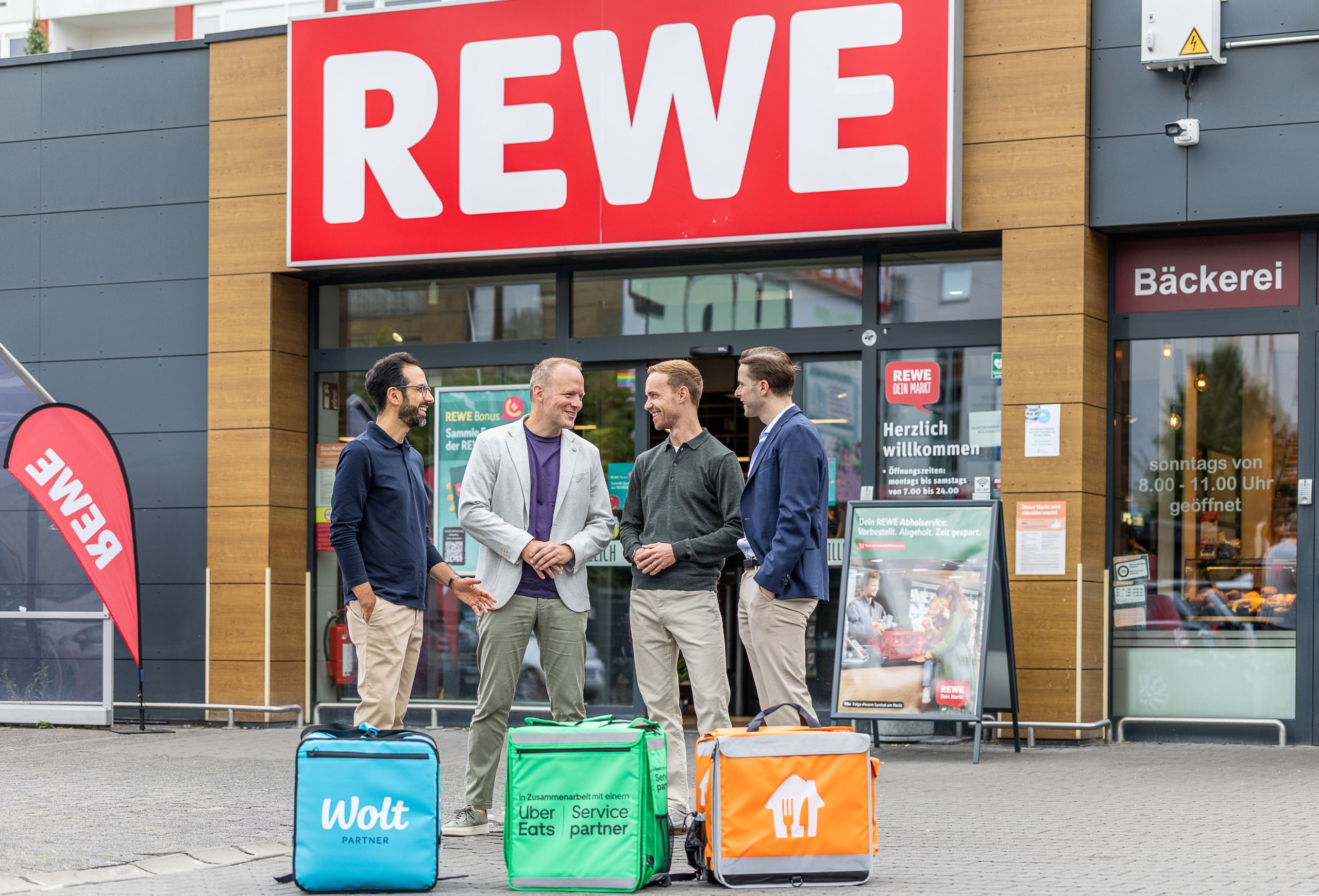 Vier Personen stehen vor dem Eingang eines REWE-Markts. Im Vordergrund sind Lieferboxen der Lieferdienste Wolt, Uber Eats und Lieferando zu sehen, die die Zusammenarbeit für die REWE Sofort-Lieferung symbolisieren.