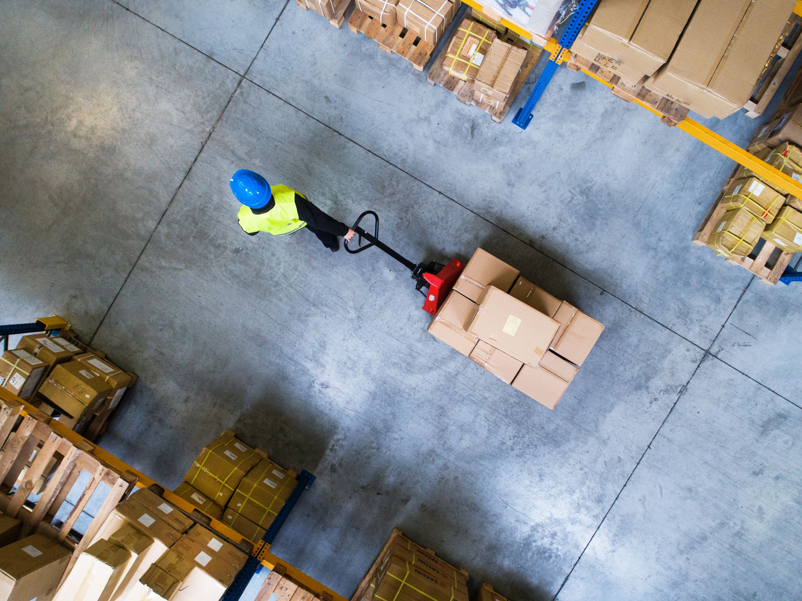 A person wearing a high-visibility vest and a hard hat is moving boxes through a warehouse using a pallet jack.