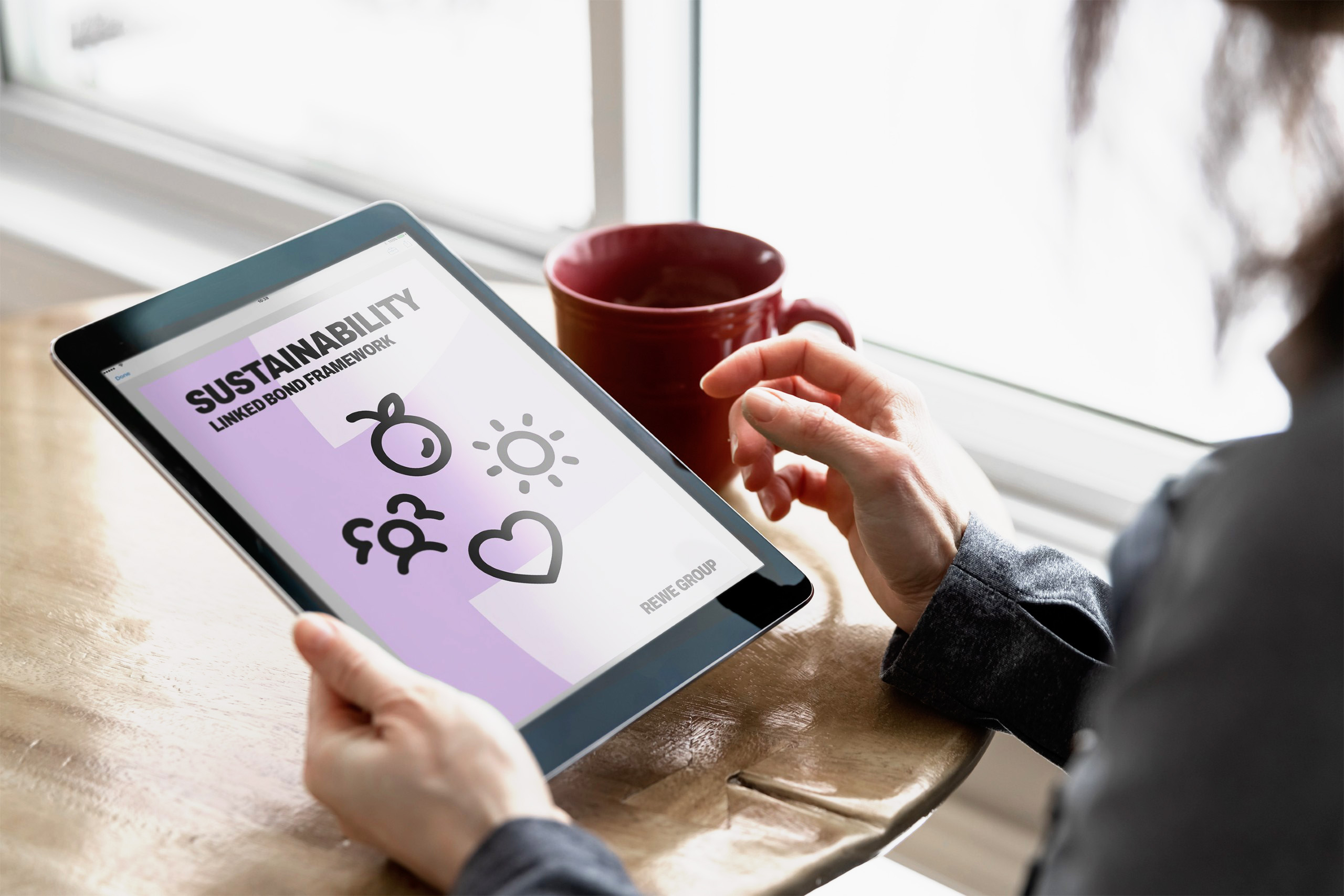 Person hält ein Tablet mit dem REWE Group Sustainability Linked Bond Framework. Auf dem Bildschirm sind Symbole für Nachhaltigkeit zu sehen.